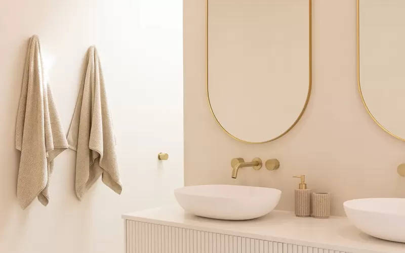 A modern bathroom with a double sink vanity, two oval mirrors with gold frames, and beige textured towels hanging on the wall