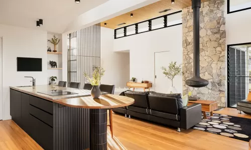 Modern open-plan living room with wooden floors, a black kitchen island with marble top, a black leather couch, a hanging fireplace, and a stone feature wall