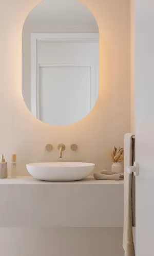 A minimalist bathroom with a round mirror, white vessel sink with golden faucet, and beige countertop with soap dispenser, small vase with dried plants, and folded towels