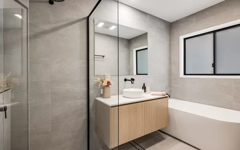 Modern bathroom interior with gray tile walls, glass shower, wooden vanity with white basin, mirror, and bathtub