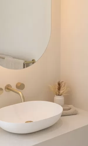 A modern bathroom with a white oval sink, gold faucet, round mirror, and decorative dried plants in a small white vase