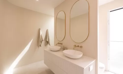 Modern minimalist bathroom with two oval mirrors, white bowl sinks on a wooden vanity, and beige towels hanging on the wall
