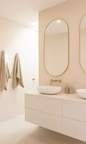 A modern bathroom with beige walls, featuring two oval-shaped mirrors with gold frames above a white vanity with ribbed drawers