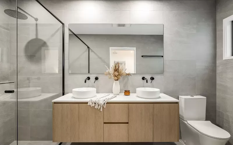 A modern bathroom with gray tiled walls, a glass shower door, white countertops with two vessel sinks
