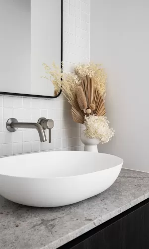 A modern bathroom vanity with a white vessel sink, a gray stone countertop, white tiles, and dried floral decor