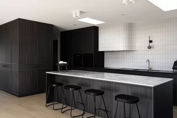 A modern kitchen with dark wood cabinetry, a large marble island, black stools, and a white tile backsplash