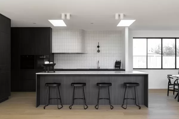 A modern kitchen with a dark wood island, black stools, white tiled backsplash, and large windows