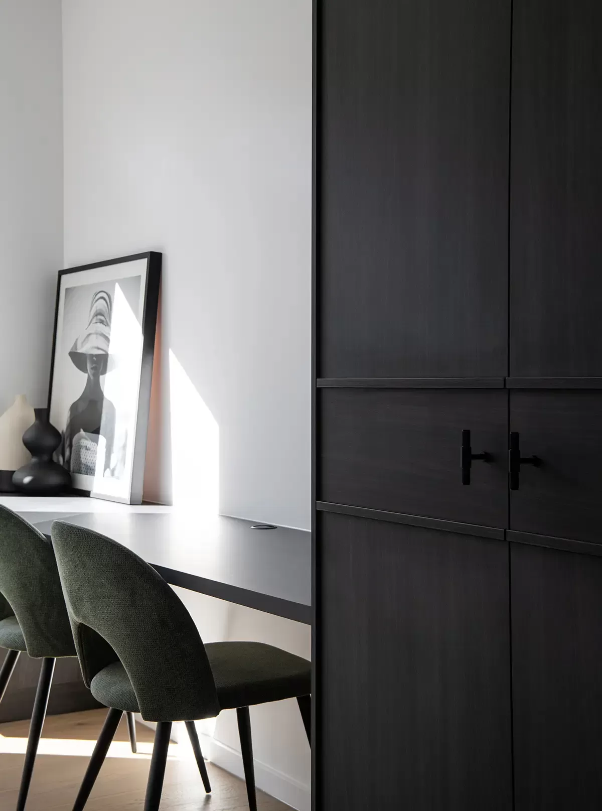 A modern home office with dark wood cabinetry, a sleek black desk, green chairs, and framed black-and-white art