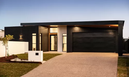 A modern house with a black brick exterior, large windows, and a spacious driveway at dusk