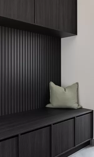 A modern mudroom with dark wood cabinetry, a black slatted wall, and a soft green cushion on the bench