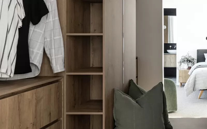 A modern walk-in closet with wood cabinetry, open shelving, hanging clothes, and a bench with green cushions