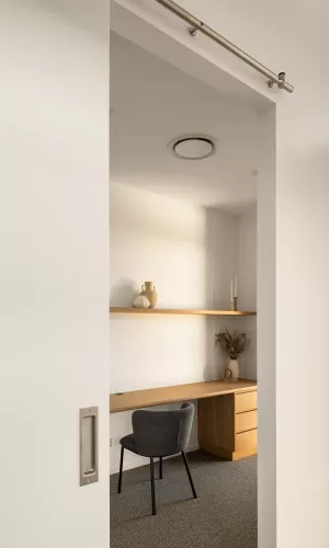 A home office with a wooden desk, grey chair, floating shelves, and minimalist decor, seen through a doorway