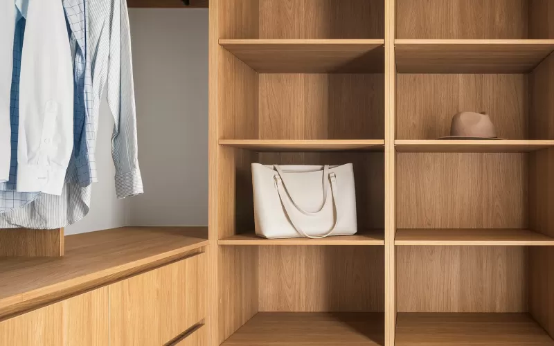 A walk-in closet with wooden shelves and drawers, containing hanging shirts, a white bag, and a beige hat