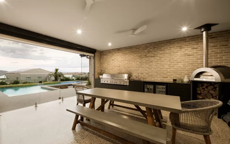 Outdoor kitchen with brick wall, barbecue grill, wood-fired oven, dining table, and pool view