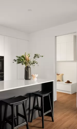 A modern kitchen with black stools, a white countertop, and a vase with green foliage, leading to a cozy room