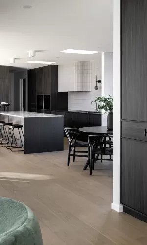 A modern kitchen with dark cabinetry, a sleek marble island, black chairs around a dining table, and light wood flooring