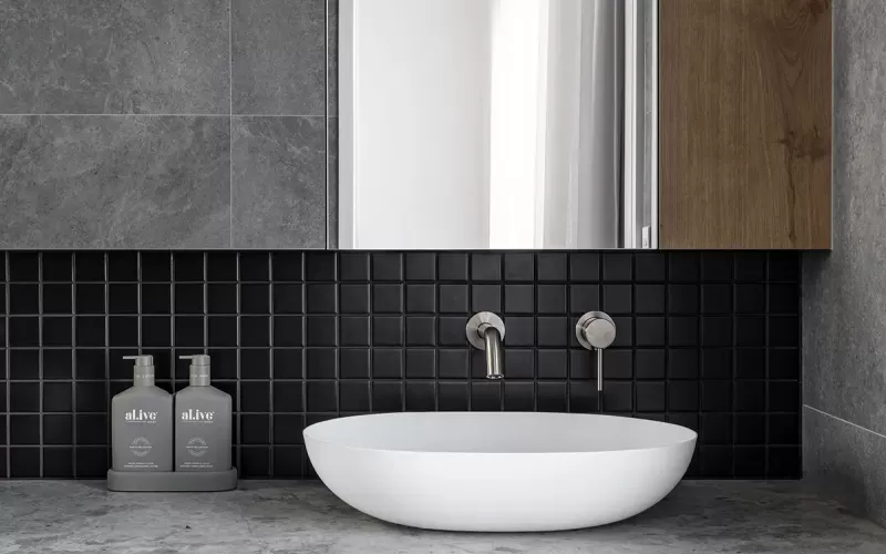 A modern bathroom with gray stone walls, a wood vanity, a white vessel sink, and black square tile backsplash