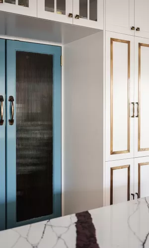 White kitchen cabinets with brass accents, blue glass-paneled doors, and marble countertop in foreground