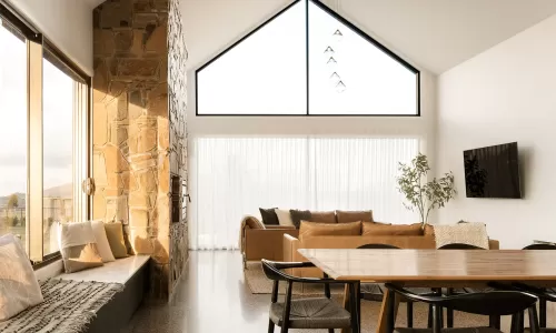 Living room with stone fireplace, vaulted ceiling, large windows, beige couch, and wooden dining table