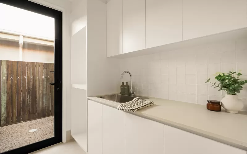 A minimalist laundry room with white cabinets, a beige countertop, a stainless steel sink, and a glass door