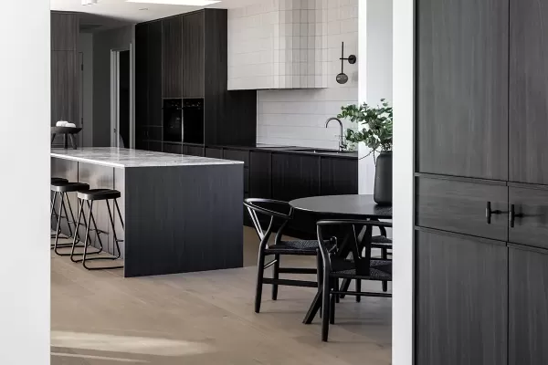 A modern kitchen and dining area with dark wood cabinetry, a marble island, black chairs, and light wood flooring